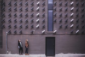 A brick wall with a lot of black and a couple of white surveillance cameras who are all watching two women in front of the building
