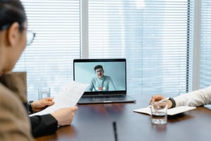 a meeting of three people two of whom sit at a table and one is only on a laptop screen present