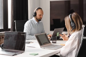two people with headsets working on laptops on opposite sides of a table in an office